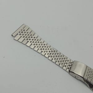 May include: A silver-toned watch band with a woven design. The band is made of small, rectangular metal links, and it has a clasp with the brand name "CASIO" engraved on it. The band is designed to attach to a watch.