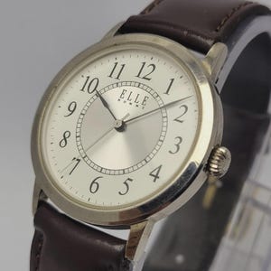 Könnte beinhalten: Silberfarbene Armbanduhr mit braunem Lederarmband. Das Zifferblatt der Uhr ist silberfarben mit schwarzen Ziffern und Zeigern. Der Markenname "ELLE HOMME" ist auf dem Zifferblatt aufgedruckt.