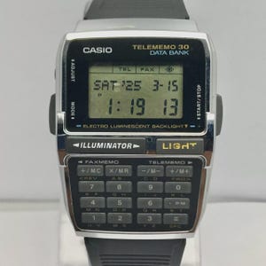 May include: A silver Casio Telememo 30 Data Bank watch with a black strap. The watch face displays the date and time, and features a calculator keypad. The watch has buttons for adjustment, mode, start/stop, and light.