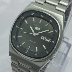 May include: A silver-toned Seiko 5 automatic wristwatch with a dark gray face. The watch features a rectangular case, a metal band, and a day-date display. The watch hands and hour markers are silver. The watch face displays the day as "SAT 12".