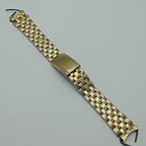 May include: A gold-toned watch band with a rectangular link design. The band features a fold-over clasp and is made of metal. The watch band is shown against a light gray background.