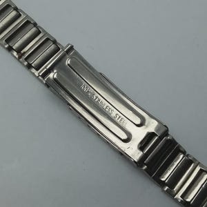 May include: Close-up of a stainless steel watch band. The band is silver and features a clasp with the text "100% STAINLESS STEEL" engraved on it. The band is made of multiple rectangular links.