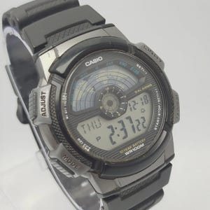 May include: A black Casio digital watch with a black rubber strap. The watch face displays the time, day, and date. Features include an alarm, stopwatch, and light. The watch is water-resistant up to 100 metres.