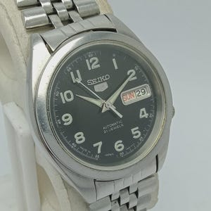 Puede incluir: Un reloj de pulsera automático Seiko 5 plateado con esfera negra. El reloj presenta una visualización de día y fecha, agujas luminosas y una pulsera de acero inoxidable. La esfera muestra el texto "SEIKO", "AUTOMATIC 21 JEWELS" y "JAPAN".