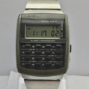 May include: A vintage Casio digital watch with a calculator keypad. The watch face displays the time as 1:17:02 AM. The watch has a silver band and a dark gray face with the Casio logo and the words "Water Resist" and "Alarm Chronograph".