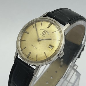 Puede incluir: Un reloj Favre-Leuba Daymatic vintage con esfera dorada, caja plateada y correa de cuero negro. La esfera del reloj muestra la fecha y el nombre de la marca. Un reloj clásico.