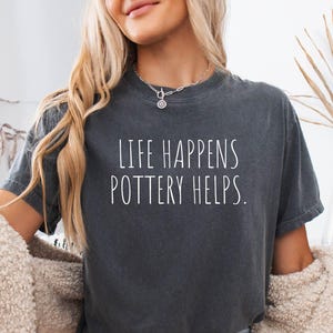 May include: A charcoal gray short-sleeve t-shirt featuring the white text "LIFE HAPPENS POTTERY HELPS." printed in a simple, clean font across the chest. The shirt has a crew neckline and a relaxed fit, paired with light blue denim shorts.