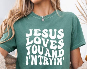 May include: A teal t-shirt with the text "JESUS LOVES YOU AND I'M TRYIN'" in white, retro-style lettering. The shirt is paired with denim shorts. The image has a fall theme, with decorative pumpkins and a woven pumpkin.