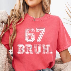 May include: A coral-colored t-shirt with the number "67" and the word "BRUH" printed in white, collegiate-style lettering. The shirt is worn with denim shorts and a cream-colored cardigan. The overall theme is casual and trendy.