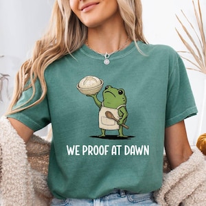 May include: Teal-colored t-shirt featuring a cartoon frog chef holding a bowl of dough and a wooden spoon. The text "WE PROOF AT DAWN" is printed in white. The shirt is paired with denim shorts.