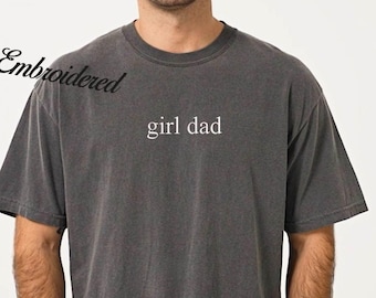 May include: Dark gray, short-sleeved t-shirt with the embroidered text "girl dad" in white. The shirt has a vintage, washed appearance and is paired with blue jeans. A watch is visible on the wrist.