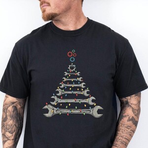 May include: Black t-shirt featuring a Christmas tree design made of wrenches, gears, and colorful string lights. The graphic is centered on the front of the shirt. The shirt is made of a soft material.