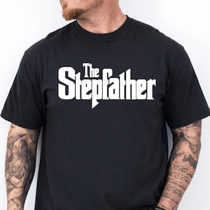 May include: Black t-shirt with the white text "The Stepfather" in a stylized font. The shirt is a crew neck and appears to be made of cotton. The person wearing the shirt is also wearing blue jeans.