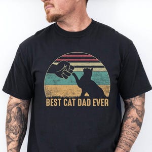 May include: Black t-shirt with a graphic design featuring a cat silhouette fist-bumping a hand, set against a retro sunset. The text "BEST CAT DAD EVER" is printed below the graphic. The shirt is made of a soft material.