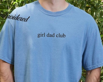 May include: A light blue t-shirt with the text "girl dad club" in black. The shirt is a crew neck style and appears to be made of a soft material. The word "embroidered" is written on the shirt in cursive.