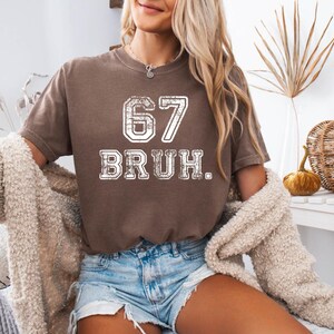 May include: A brown t-shirt with the number "67" and the word "BRUH" in a distressed white font. The shirt is paired with denim shorts. A cream-colored sweater is draped over the shoulders.