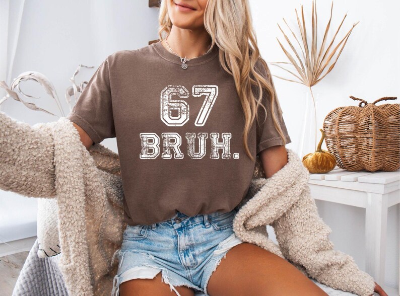 May include: A brown t-shirt with the number "67" and the word "BRUH" in a distressed white font. The shirt is paired with denim shorts. A cream-colored sweater is draped over the shoulders.