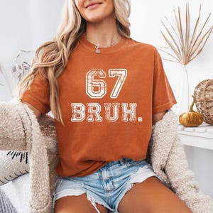 May include: Burnt orange t-shirt with the number "67" and the word "BRUH" in white, distressed lettering. The shirt is worn with denim shorts and a cream-colored cardigan. The overall theme is casual and trendy.