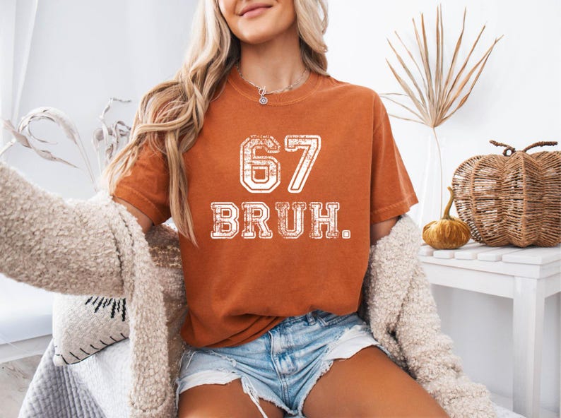 May include: Burnt orange t-shirt with the number "67" and the word "BRUH" in white, distressed lettering. The shirt is worn with denim shorts and a cream-colored cardigan. The overall theme is casual and trendy.