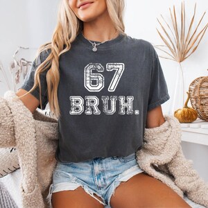 May include: Dark gray t-shirt with the number "67" and the word "BRUH" in white, distressed lettering. The shirt is worn with denim shorts and a cream-colored cardigan. The overall aesthetic is casual and relaxed.