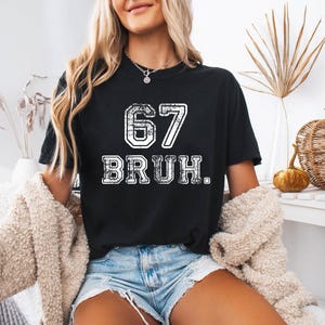 May include: Black t-shirt with the number "67" and the word "BRUH" in white, distressed lettering. The shirt is a crew neck style. The image shows a person wearing the shirt.