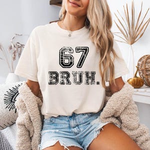 May include: Cream-colored t-shirt with the number "67" and the word "BRUH" printed in black, distressed lettering. The shirt is a casual style, suitable for everyday wear.