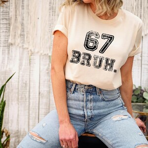 May include: A cream-colored t-shirt with the number "67" and the word "BRUH" printed in a distressed black font. The shirt is paired with light blue ripped jeans.