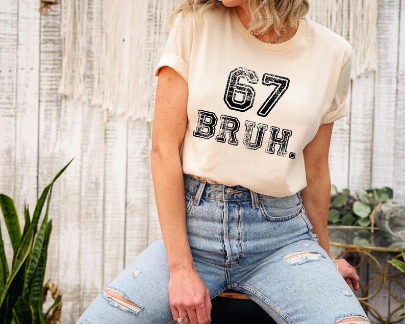 May include: A cream-colored t-shirt with the number "67" and the word "BRUH" printed in a distressed black font. The shirt is paired with light blue ripped jeans.