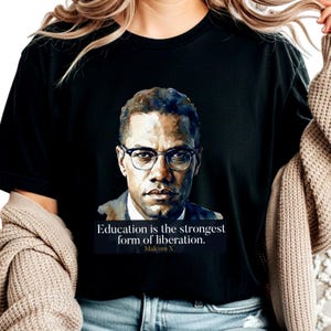 Może przedstawiać: Czarny t-shirt z portretem Malcolma X w okularach. Obrazowi towarzyszy cytat "Edukacja jest najsilniejszą formą wyzwolenia. Malcolm X" w białym tekście. Koszulka ma okrągły dekolt.