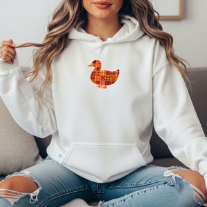 May include: White hoodie with a front pocket and a duck graphic. The duck is made of a patchwork design in shades of orange, red, and yellow. The hoodie is made of a soft material and has a drawstring hood.