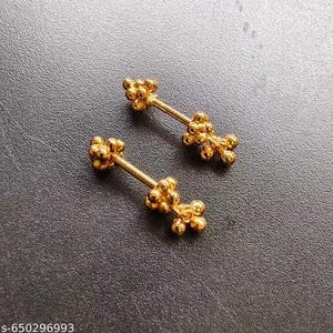 Peut inclure: Deux boucles d'oreilles dorées en forme d'haltères. Chaque boucle d'oreille comporte une barre centrale avec des accents en forme de boules groupées à chaque extrémité et au milieu. Les boucles d'oreilles sont présentées sur une surface gris foncé.