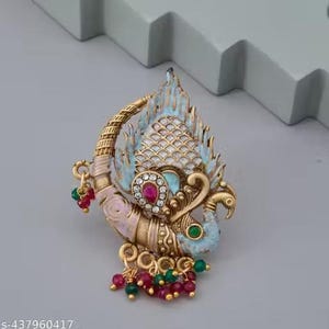 May include: Ornate gold-toned peacock brooch with intricate detailing. The brooch features a central cluster of red and clear gemstones, with accents of light blue and pink enamel. Small red and green beads dangle from the bottom.