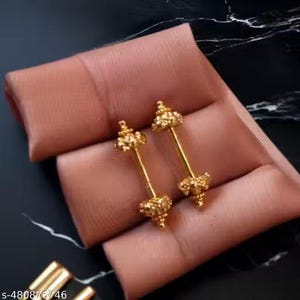 May include: Two gold-colored barbell earrings with ornate, textured ends. The earrings are displayed on a peach-colored surface against a dark, marbled background. The earrings have a symmetrical design.