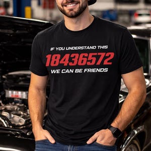 Op de afbeelding: Zwart t-shirt met de tekst "IF YOU UNDERSTAND THIS 18436572 WE CAN BE FRIENDS" in rood en wit. Het t-shirt wordt gedragen door een persoon die een zwarte pet draagt en voor een auto staat.
