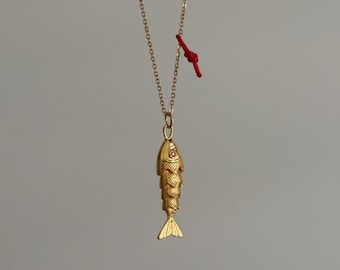 Puede incluir: Collar con colgante de pez dorado. El colgante de pez tiene escamas detalladas y un pequeño acento rojo. La cadena del collar es delicada y un cordón rojo está unido cerca del cierre. El fondo es gris liso.