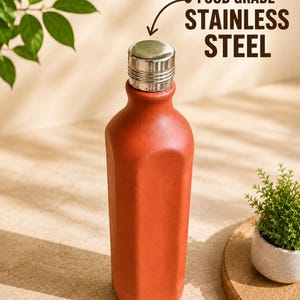 May include: A terracotta-colored bottle with a square-shaped body and a metallic silver cap. The text FOOD GRADE STAINLESS STEEL is printed near the top, with an arrow pointing to the cap, indicating the material of the bottle closure.