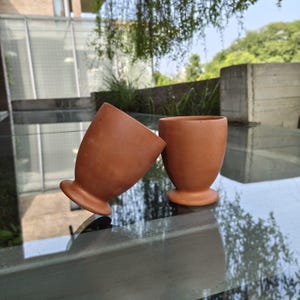 Può includere: Due tazze fatte a mano color terracotta, con base arrotondata e parte superiore svasata. Una tazza è inclinata, rivelando l'interno. Le tazze sono su una superficie riflettente, con uno sfondo esterno sfocato.