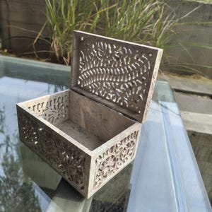 May include: Open, rectangular, beige stone box with intricate floral and leaf carvings. The lid is open, revealing the empty interior. The box is sitting on a glass surface, with greenery in the background. A decorative storage item.