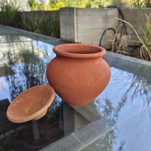 Puede incluir: Una olla de arcilla de terracota con su tapa. La olla es redonda con una abertura ancha y una superficie ligeramente texturizada. La tapa tiene forma de plato poco profundo. Los artículos son de color marrón rojizo cálido.