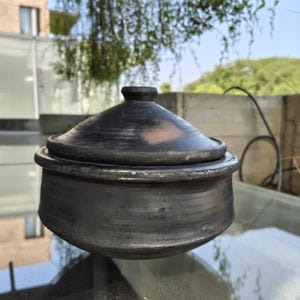 May include: A dark grey, handmade ceramic pot with a matching lid. The pot has a rounded base and a slightly flared rim. The lid has a small knob on top. The pot is sitting on a glass surface.