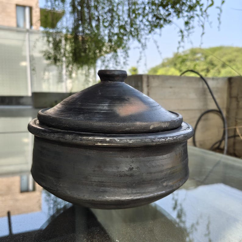 May include: A dark grey, handmade ceramic pot with a matching lid. The pot has a rounded base and a slightly flared rim. The lid has a small knob on top. The pot is sitting on a glass surface.
