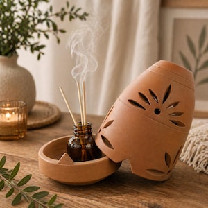 May include: A terracotta essential oil diffuser with a decorative floral design. The diffuser includes a small amber glass bottle with wooden reeds and a matching bowl. Wisps of vapor rise from the reeds, suggesting the diffusion of fragrance.