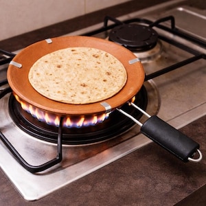 Puede incluir: Un calentador de tortillas redondo de terracota marrón con un mango negro, que sostiene una tortilla cocida sobre un quemador de estufa de gas. El quemador está encendido con llamas azules y amarillas.