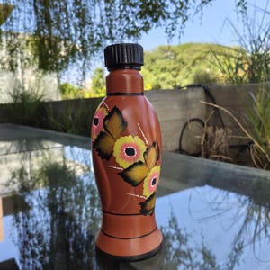 Peut inclure: Une bouteille décorative marron avec un bouchon noir. La bouteille est ornée de motifs floraux peints à la main en jaune, rose et noir. La bouteille est posée sur une surface en verre.