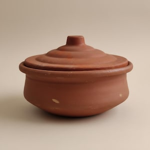 Puede incluir: Una olla de cocina de terracota con su tapa a juego. La olla es de color marrón rojizo cálido y tiene una forma redondeada con un borde ligeramente ensanchado. La tapa tiene un pomo y anillos concéntricos.