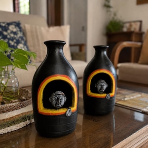 May include: Two black ceramic vases with a matte finish, each featuring a central arched window cutout. Inside each window, a small grey sculpted figure with a round head and one hand rests on the ledge. The arch is outlined in yellow and orange.