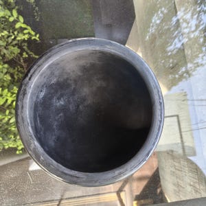 May include: A dark grey, round ceramic bowl with a wide rim. The bowl has a textured surface and appears to be empty. The bowl is sitting on a reflective surface.