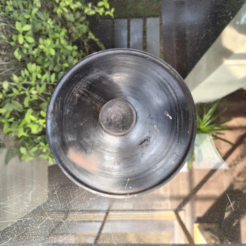 May include: A top-down view of a black, round, ceramic pot with a small, circular knob in the centre. The pot has a slightly textured surface and appears handmade. The pot is sitting on a glass surface.
