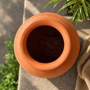 May include: A top-down view of a terracotta pot featuring a wide, circular opening and a smooth, rounded rim. The vessel is crafted from natural clay, displaying a warm, earthy orange-brown color with a matte, unglazed finish and a deep interior.