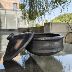May include: A dark grey, handmade clay pot with a matching lid. The pot is round with a wide opening and a slightly textured surface. The lid is oval-shaped with a small handle. The pot and lid are on a reflective surface.